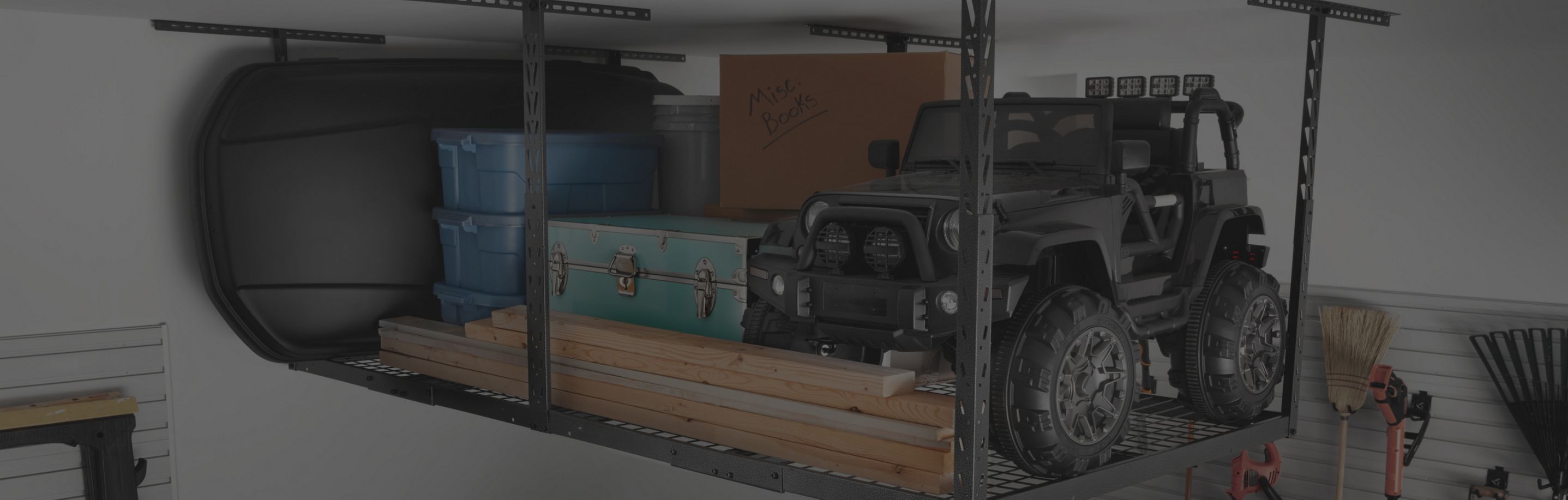 Gladiator Overhead Storage with blue bins, lumber, boxes, a green chest and a motorized car. Hanging on the wall is a broom, rake and gardening equipment