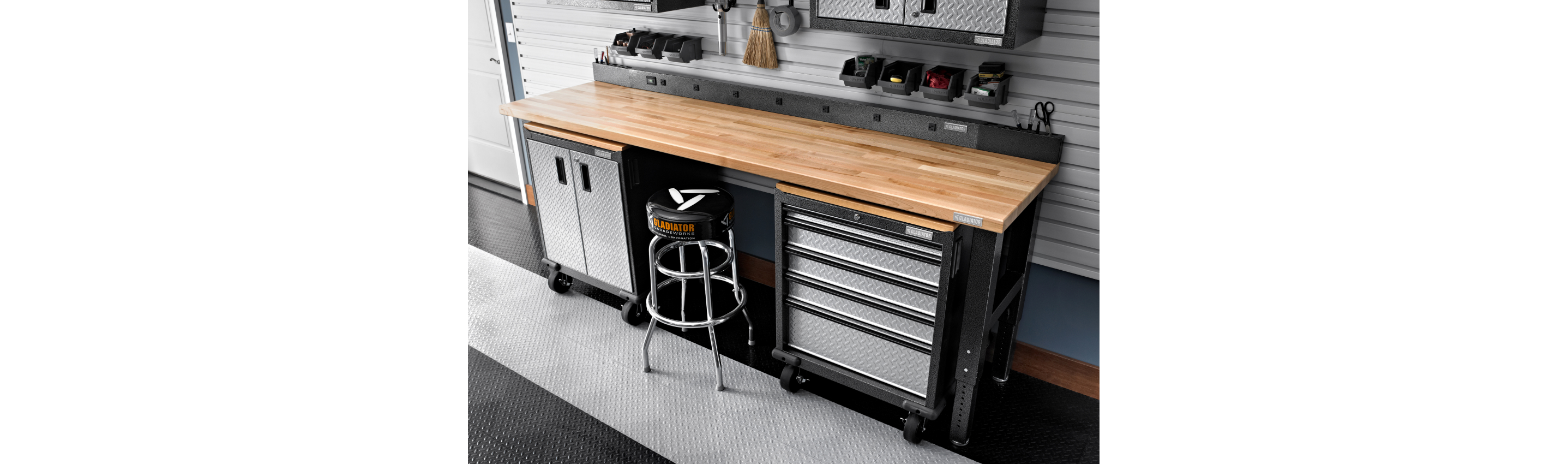 A Gladiator Workbench on two Gladiator Modular Units with a stool. Surrounding the workbench are bins, a broom, scissors and more.