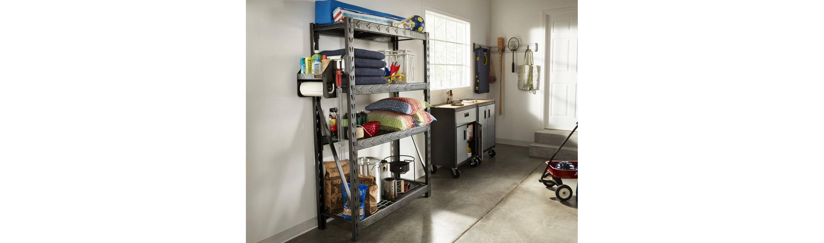 A garage with a Gladiator Shelving Unit with pillows, watering cans, a Gladiator Clean-up Caddy and other items. There's also a red wagon, a Gladiator Workbench, and items on a Gladiator GearTrack.