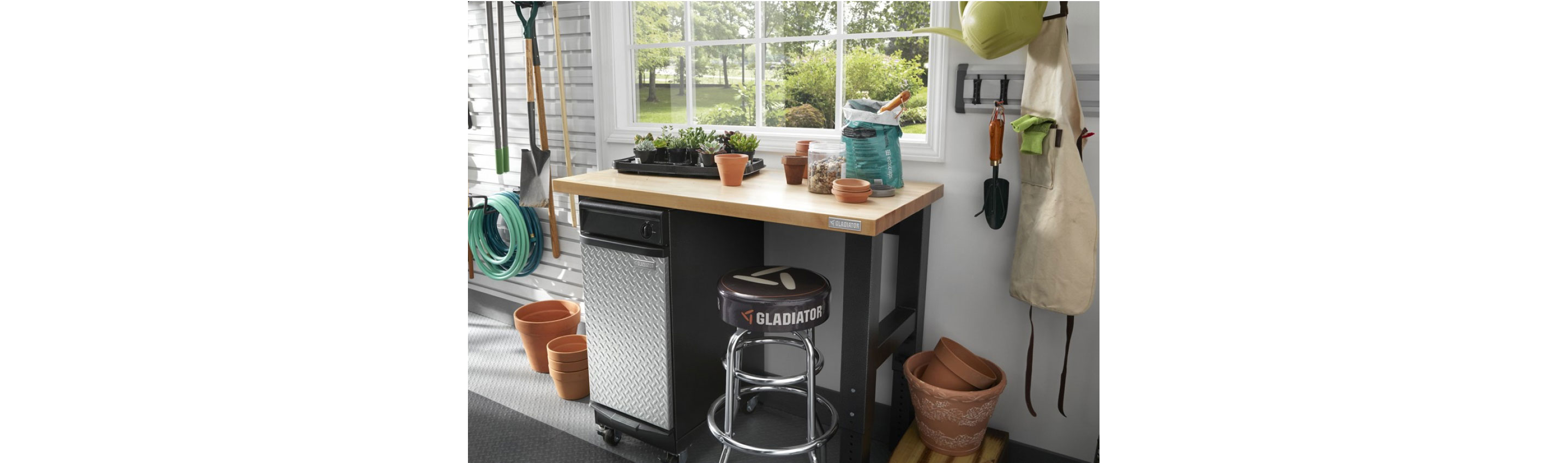 A garage with a Gladiator Workbench with a Gladiator Modular Cabinet underneath. There's a stool and several plant pots nearby. On the wall on a Gladiator GearTrack is a gardening shovel and apron. On the far wall hangs a garden hose and gardening tools