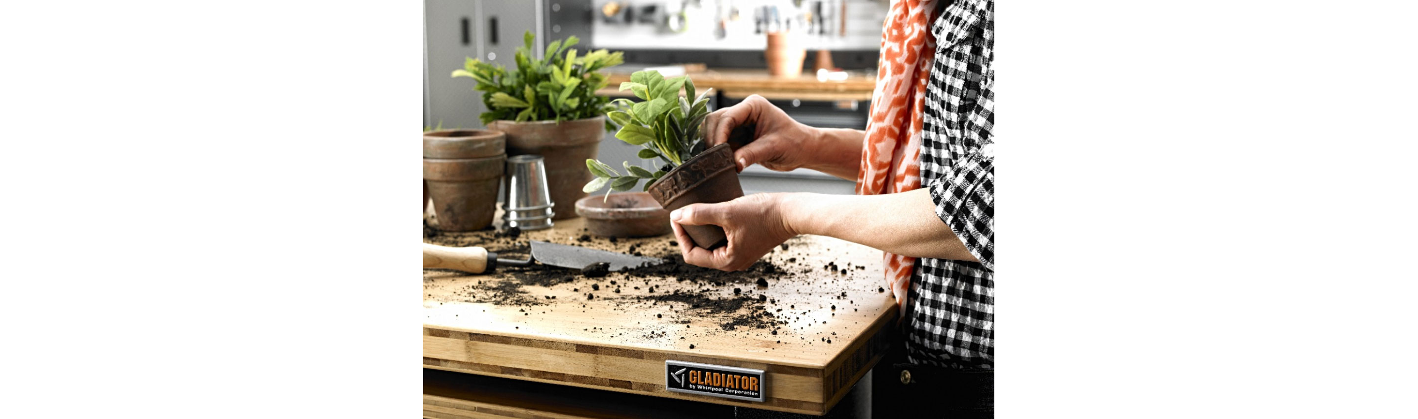 Someone in a black and white checkered shirt touches a potted plant in front of a Gladiator Workbench that's covered in soil. On the workbench are a stack of pots and a potted plant and a garden tool