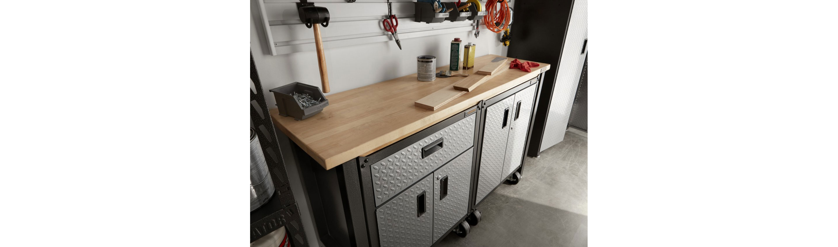 A Gladiator Workbench and Gladiator Full-Size Cabinet. On the workbench are wood planks, a Gladiator bin with nails, a rag and cans. Hanging on the wall are a mallet, scissors, wrench and extension cord